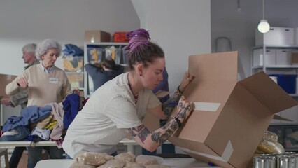 Wall Mural - Young woman with pink hair and tattoos sign cardboard box and pack food in volunteering center