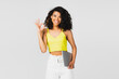 positive african american woman in yellow crop top holding laptop and showing ok isolated on grey.