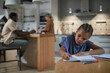 © Seventyfour - Portrait of cute black girl studying at kitchen table with multiethnic parents in background, copy space
