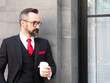 © panyajampatong - Portrait of a bearded hipster businessman in a suit standing happily drinking coffee in a business district.