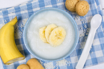 Wall Mural - Rice porridge for the baby from ground cereals in a blue bowl with a banana, cookies and a spoon on a cloth napkin. Baby's first complementary food, baby nutrition.