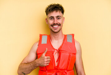 Wall Mural - Young caucasian man wearing a life jacket isolated on yellow background smiling and raising thumb up