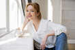 © LIGHTFIELD STUDIOS - Portrait of smiling blonde woman looking at camera near windowsill at home.