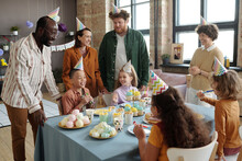 Group Of Adults In Party Hats Watching For Children While They Sitting At Table And Eating Dessert During Birthday Party