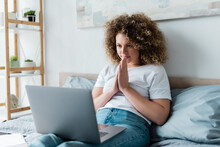 Positive Woman Sitting With Praying Hands Near Laptop In Bedroom.