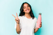 Young hispanic woman holding thermo isolated on blue background joyful and carefree showing a peace symbol with fingers.
