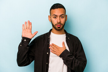 Wall Mural - Young hispanic man isolated on blue background taking an oath, putting hand on chest.