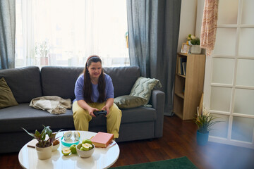 Brunette girl in casualwear sitting on couch in living room at leisure and playing video game while pressing buttons on joystick