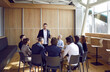 © Studio Romantic - Young business coach talking to team of people. Group of entrepreneurs having meeting with friendly business trainer, sitting in circle in modern office interior and listening to interesting story