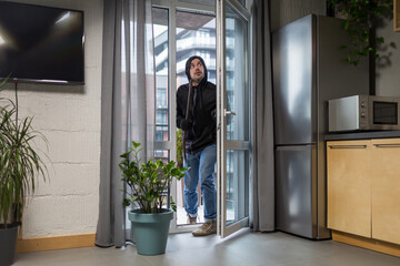 Young male robber in dark clothes carefully and quietly enters through the balcony window into the apartment, house. Holds scrap metal, fittings.