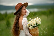 A Middle-aged Woman In A White Dress And Brown Hat Stands On A Green Field And Holds A Basket In Her Hands With A Large Bouquet Of Daisies. In The Background There Are Mountains And A Lake.