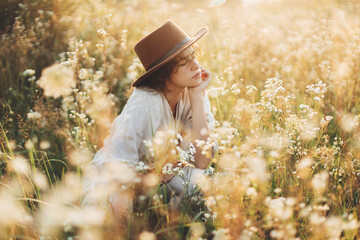 Wall Mural - Stylish boho woman in hat sitting among wildflowers in warm sunset light. Summer delight and travel. Young female in rustic linen cloth relaxing in summer meadow. Atmospheric moment