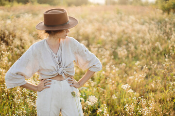 Wall Mural - Stylish boho woman in hat posing among wildflowers in sunset light. Summer delight and travel. Young female in rustic linen cloth relaxing in summer meadow. Atmospheric moment