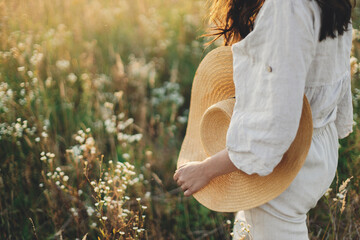 Poster - Stylish boho woman walking with straw hat in hand close up among wildflowers in sunset light. Atmospheric moment. Summer travel. Young female in rustic linen cloth relaxing in summer meadow