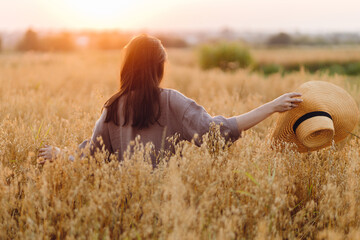 Wall Mural - Stylish woman with straw hat standing in oat field in sunset light. Atmospheric tranquil moment. Young female in rustic linen dress relaxing in evening summer countryside. Rural slow life