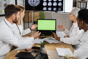 Teamwork of four multiethnic doctors wearing white lab coats and pointing on laptop with green screen while working in polyclinic boardroom. Concept of effective team cooperation in healthcare field.