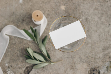 Blank business card mockup on champagne glass with golden rim. Blurred concrete ground with olive tree branch. Mediterranean summer wedding stationery, invitation. Birthday party celebration.