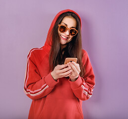 Canvas Print - Happy smiling brunette woman of caucasian holding in hand and use mobile cell phone in fashion red hoodie and sunglasses on purple bright background with empty copy space. Studio