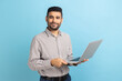 Smiling man freelancer with beard standing and holding laptop, satisfied with teleworking, likes his job, looking at camera, wearing striped shirt. Indoor studio shot isolated on blue background.