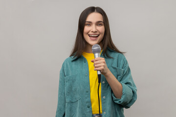 Portrait of positive woman reporter standing with microphone in hands, sing songs with happy expression or telling news, wearing casual style jacket. Indoor studio shot isolated on gray background.