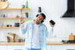 © kucherav - Happy African American man in headphones and with mobile phone listening to your favorite track, dancing in home kitchen