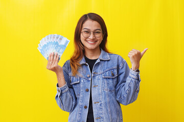 Pretty young asian woman standing while holding an Indonesian money and pointing something.