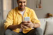 © Prostock-studio - Young African American man sitting on sofa with smartphone, working or studying online, browsing social network at home