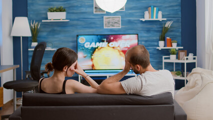 Sticker - Couple disappointed because of losing online console space shooter while playing using wireless controller while sitting on couch. Young woman and boyfriend arguing over failed level in action game.