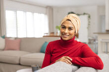 Portrait Of Smiling Biracial Young Woman In Headscarf Sitting On Sofa At Home