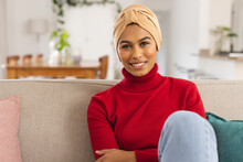 Portrait Of Happy Biracial Young Woman Wearing Headscarf Sitting On Sofa At Home
