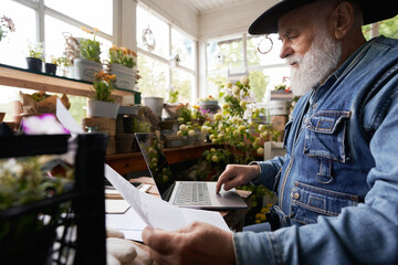 Aged man working with laptop and documents in flower shop