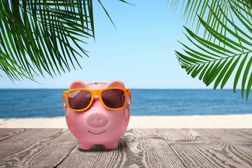 Canvas Print - Saving money for summer vacation. Piggy bank with sunglasses on wooden surface near sandy beach and sea