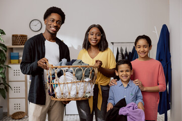 The happy cheerful family does household chores together in the laundry room. Parents hold a large wicker basket with clothes to sort, young children daughter son help learn to clean.