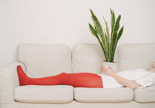 Girl In Red Tights Lies On A Beige Sofa With Houseplant On Her Stomach. Waiting Is Tired. Artistic Creative Conceptual  Composition. Horizontal. Restrained Emotions In  Appointment With Psychotherapis
