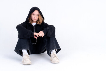 A caucasian teenage girl in a black hoodie with a hood is sitting on the floor on a white background. Studio photo. Difficult adolescence transitional age concept