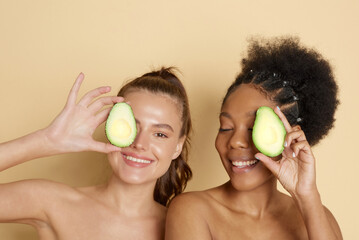 Wall Mural - Young smiling afro american and caucasian women holding an avocado in front of their eyes on a beige background.