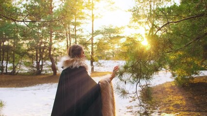 Wall Mural - Fantasy woman walks in winter forest hand touches strokes branch pine tree. Princess girl. Green long vintage cape dress. Mystic gohic lady back rear view in magic divine sun light. Nature woods snow