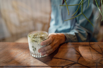 Wall Mural - Hand on a glass of Iced matcha latte green tea with milk on a wooden bar over a cafe glass window reflex at a Cafe coffee shop. Cold brew refreshment summer drink with copy and space. Selective focus