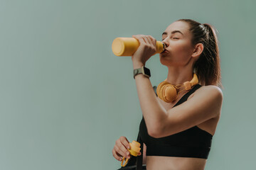 Young sporty woman in sportswear with wireless headset drinking from bottle over green background, copy space