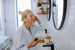 Beautiful senior woman in bathrobe applying natural face cream in bathroom, skin care and morning routine concept.