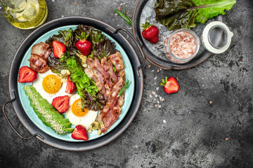 Wall Mural - Two eggs, bacon, avocado, strawberries and fresh salad for healthy breakfast on a dark background. Healthy fats, clean eating for weight loss. place for text, top view