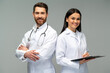 Woman doctor holding clipboard and posing with her male colleague with stethoscope with happy faces while standing isolated on grey background