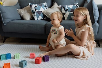 Wall Mural - Little caucasian girl 7 years old and her baby sister 1 year old. Toddler kid playing with sibling at home in living room. Family with two children at home. Love, trust, sisterhood concept