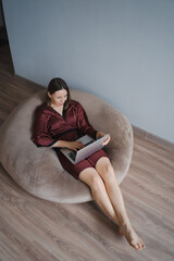 Wall Mural - Woman wearing a silk robe using laptop at home relaxing, sitting on a beanbag