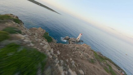 Wall Mural - FPV video, view from above, stunning aerial view from an FPV drone flying at high speed over a rocky coastline with a lighthouse illuminated during a dramatic sunset. Faro di Capo Ferro, Sardinia.
