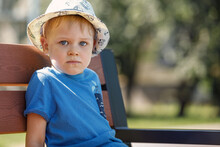Portrait Of A Cute Boy With A White Hat And A Blue Shirt. A Child Sits On A Nat Bench In The Park And Looks Straight At The Camera.