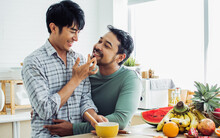 Gay LGBT Sweet Asian Couple Wearing Pajamas, Smiling With Happiness And Love, Eating, Feeding Orange, Healthy Fruits For Breakfast In Kitchen At Home In The Morning With Sunlight. Lifestyle Concept.