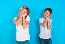 Relax And Sleep Time. Tired Two Kids Boy And Girl Standing Over Blue Studio Background With Closed Eyes Leaning On Palms Making Sleeping Gesture.