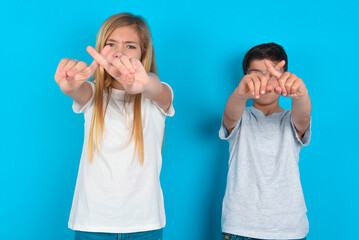 two kids boy and girl standing over blue studio background has rejection angry expression crossing fingers doing negative sign.