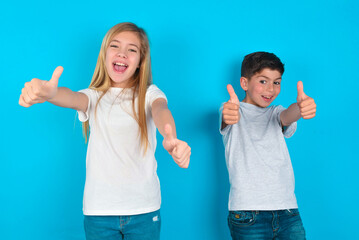 two kids boy and girl standing over blue studio background making positive gesture with thumbs up smiling and happy for success. Looking at the camera, winner gesture.